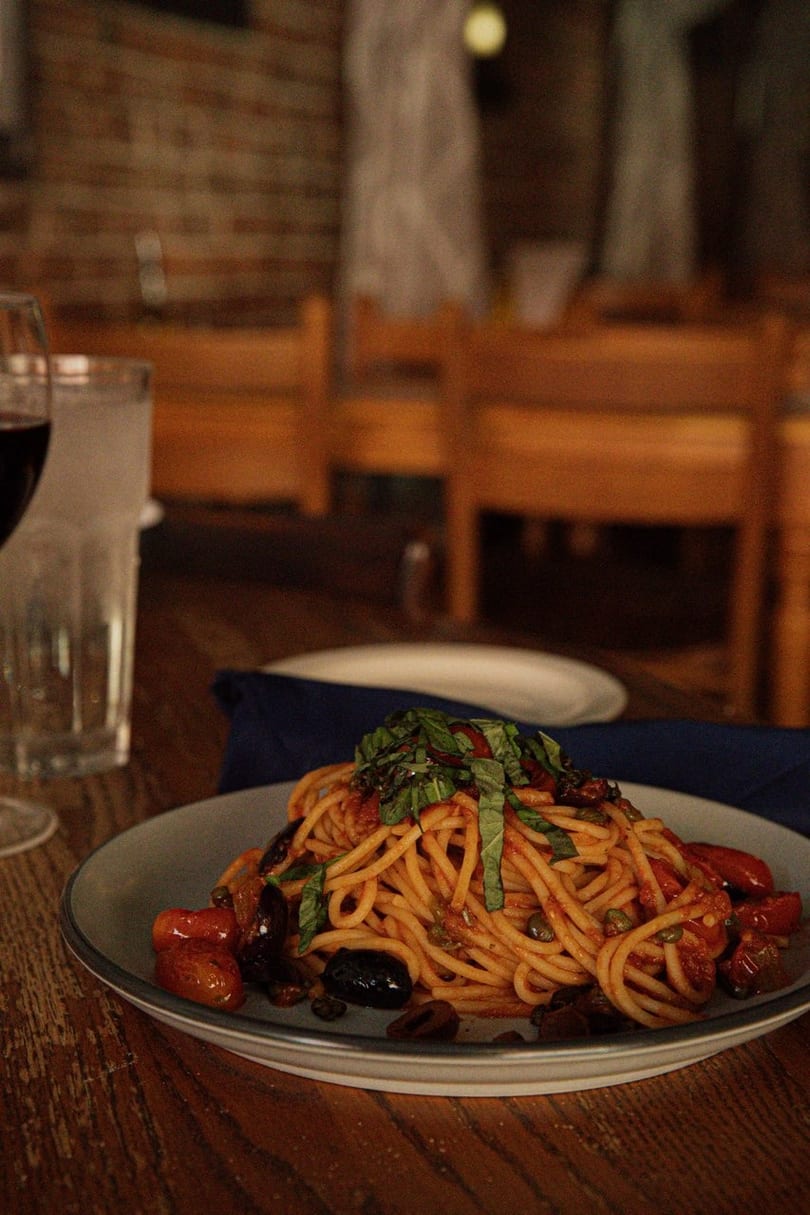 Spaghetti with tomato sauce, olives, capers, and basil on a wooden table with drinks.