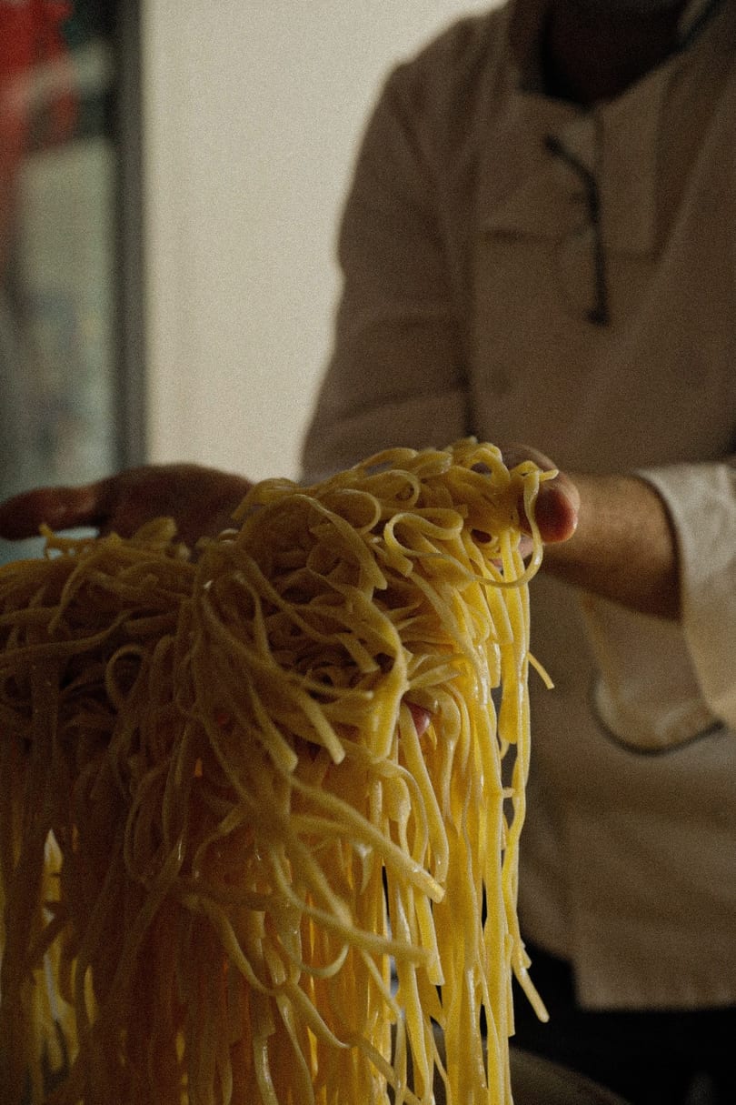 A chef's hands holding up a large, glistening tangle of freshly made yellow pasta noodles.